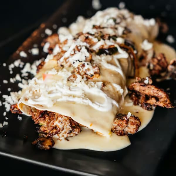 A close-up of a Mexican enchilada covered in white sauce, crumbled cheese, and meat on a black plate.