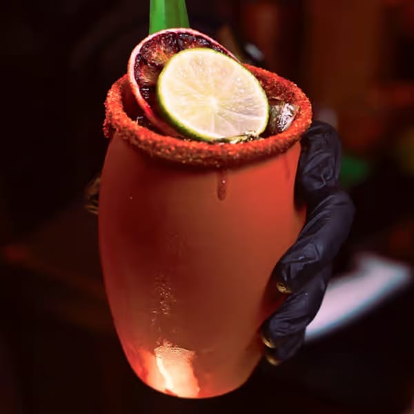 A hand in a black glove holds a red Santo Tequila cocktail garnished with lime and orange.